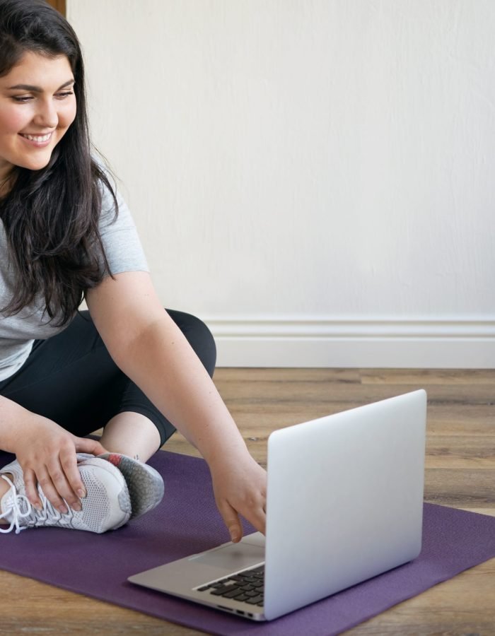 Attractive cute girl with curvy body sitting on mat exercising to lose weight using laptop, doing distant training via video conference chat under supervision of professional fitness instructor