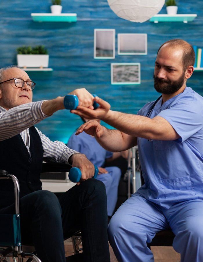 Practitioner man doctor helping retiree senior male in wheelchair to do physiotherapy strength exercise using dumbbell. Social nursing services at home. Healthcare recovery. Support worker helper