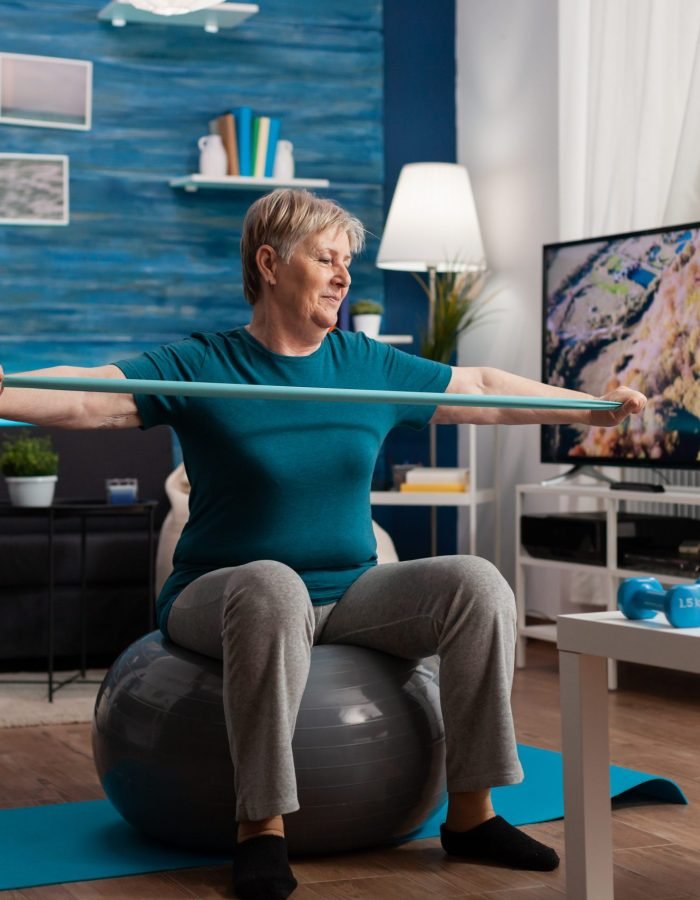 Retired senior man sitting on fitness swiss ball in living room doing wellness fitness workout streching arm muscles using aerobics elastic band. Pensioner training body resistance in living room