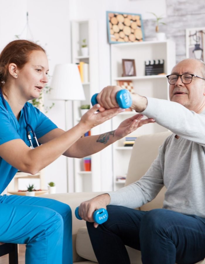 Senior man in nursing home with doing physical therapy with help from nurse using dumbbells.